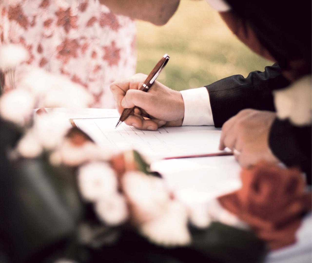 A hand signing the marriage register with a fountain pen, a bouquet of white and pink roses beside it